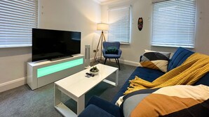 A modern living room with a flat-screen TV, a white coffee table, a blue armchair, and a sofa with colorful cushions.