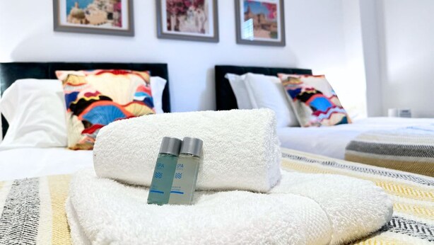 A neatly made bed with a white towel and two bottles of SPA products, framed pictures on the wall, and a patterned bedspread.