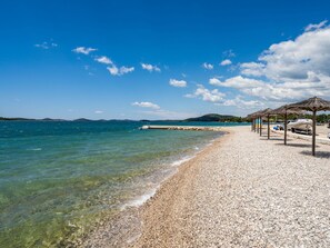 A pebble beach with clear water, a pier, and a row of thatched umbrellas. - Wonderful private villa with private pool, A/C, WIFI, TV, terrace, pets allowed and panoramic view (Biograd Na Moru)
