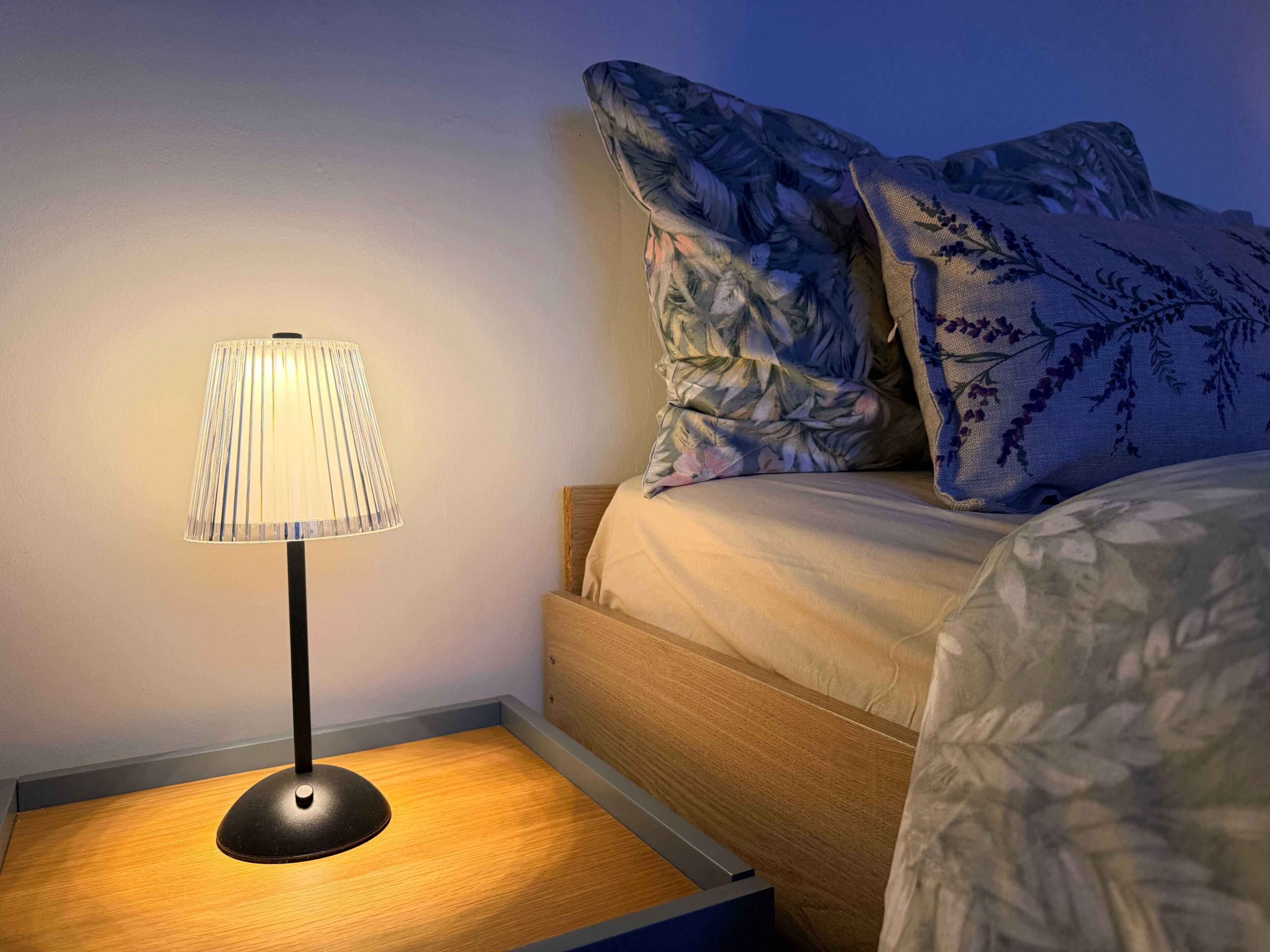 A bedside table with a lamp, a bed with floral-patterned bedding, and a nightstand.