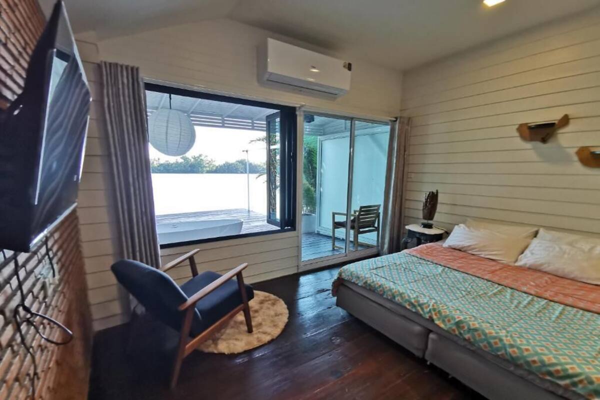A bedroom with a bed, a chair, a TV, and a sliding glass door leading to a balcony.