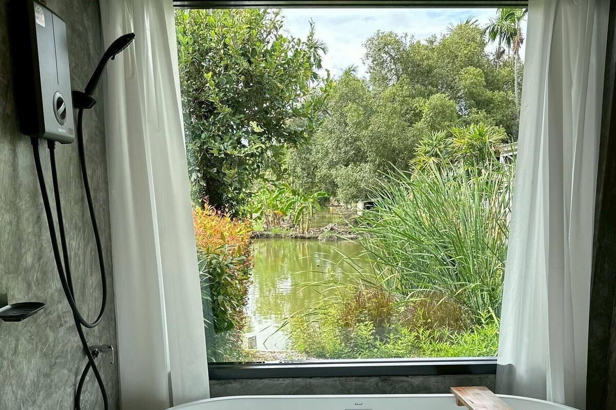 A modern bathroom with a freestanding bathtub, a window with curtains, and a view of a pond and greenery.