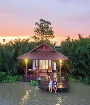 A wooden house with a porch, surrounded by water and palm trees, at sunset. - House of moon phase (Chachoengsao)