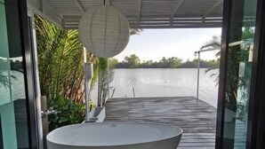 A modern bathroom with a large white bathtub, a hanging paper lantern, and a view of a body of water through open doors. - House of moon phase (Chachoengsao)