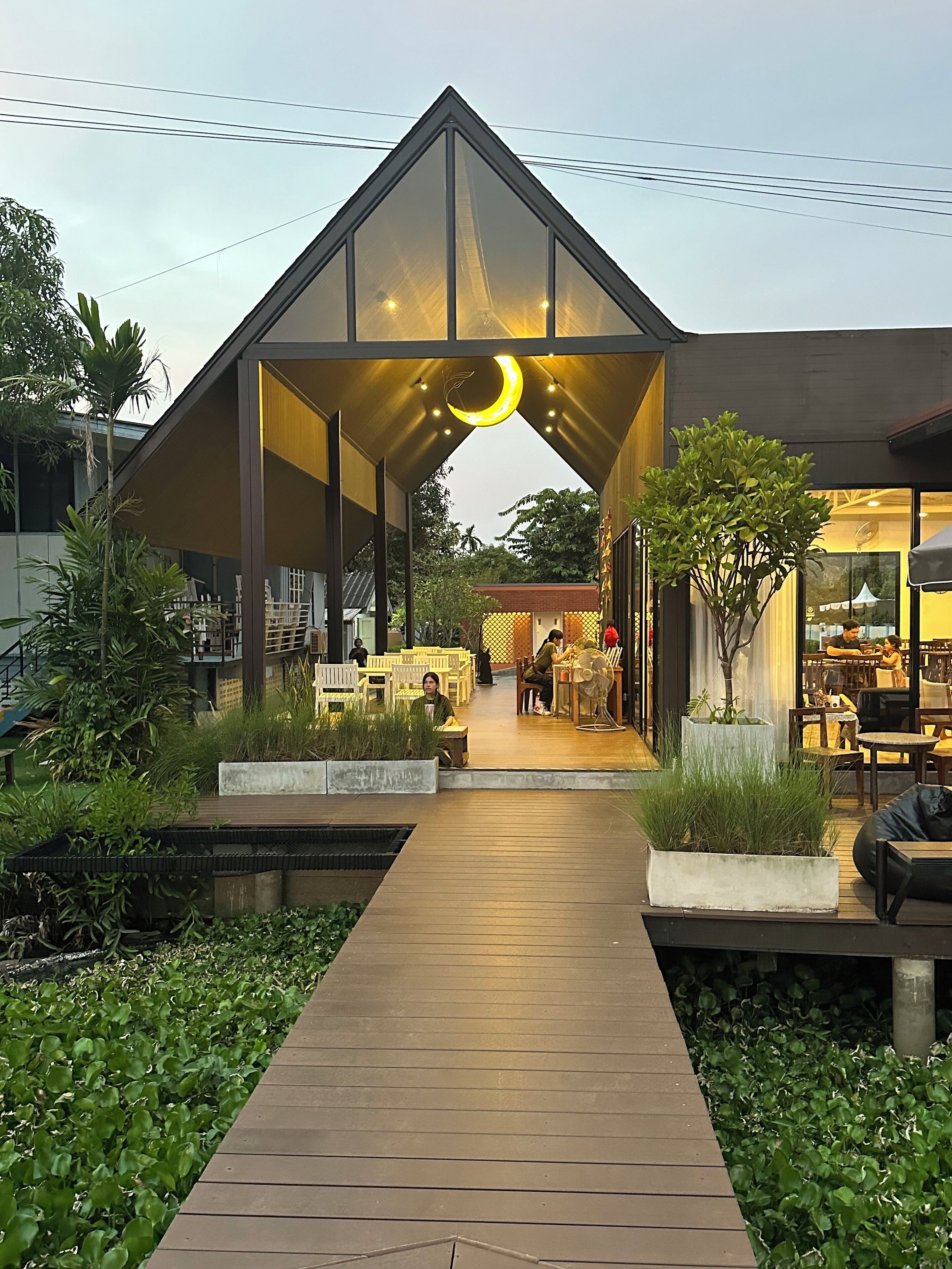 A modern building with a triangular roof structure, a wooden pathway leading to an outdoor seating area, and potted plants.