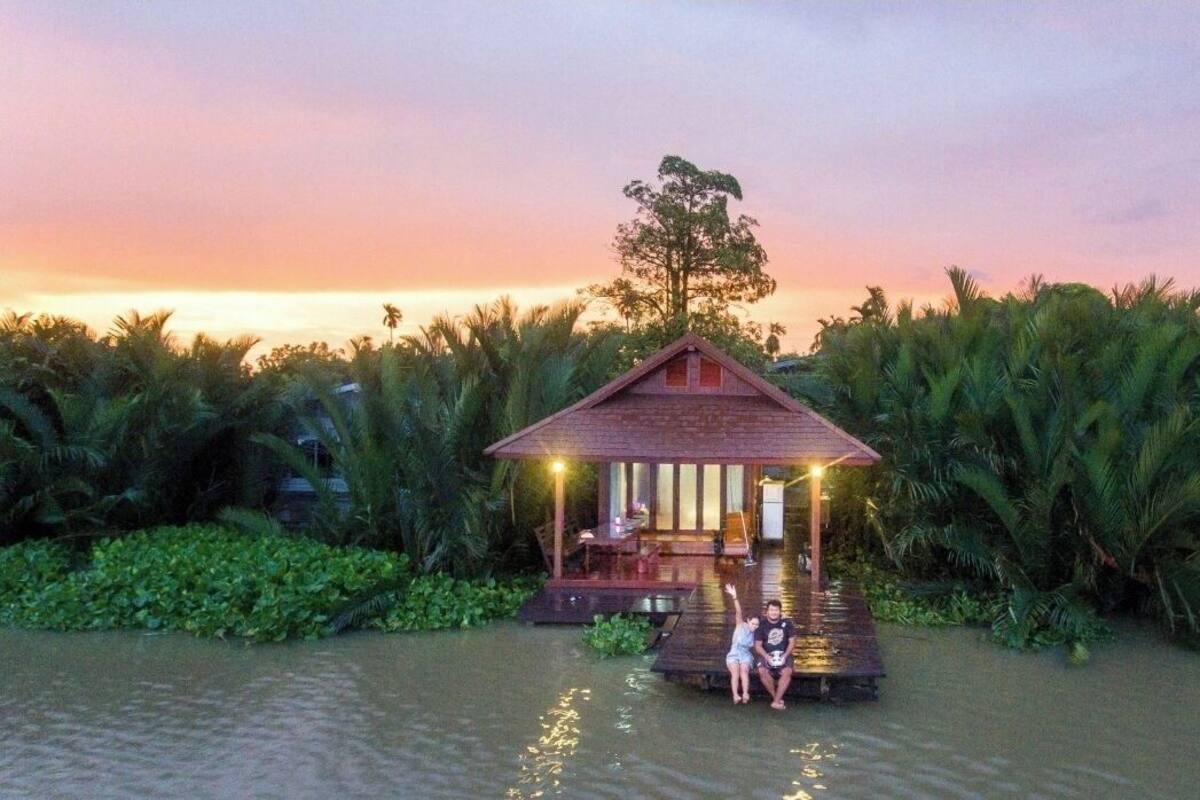 A wooden structure with a thatched roof, surrounded by water and palm trees, at dusk.
