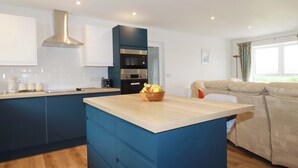 A modern kitchen with a central island, a built-in oven, and a dining area with a sofa. - Folly View (Redruth)