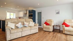 A living room with a beige sofa, two armchairs, a wooden floor, and a kitchen in the background. - Folly View (Redruth)