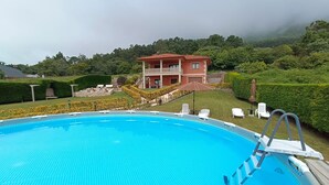 A swimming pool with a clear blue surface, surrounded by a well-maintained lawn and a two-story building with a red roof in the background. - OASIS INN PEDORNES.A retreat between sea & mountains on Galicia’s Atlantic coast (Pedornes)