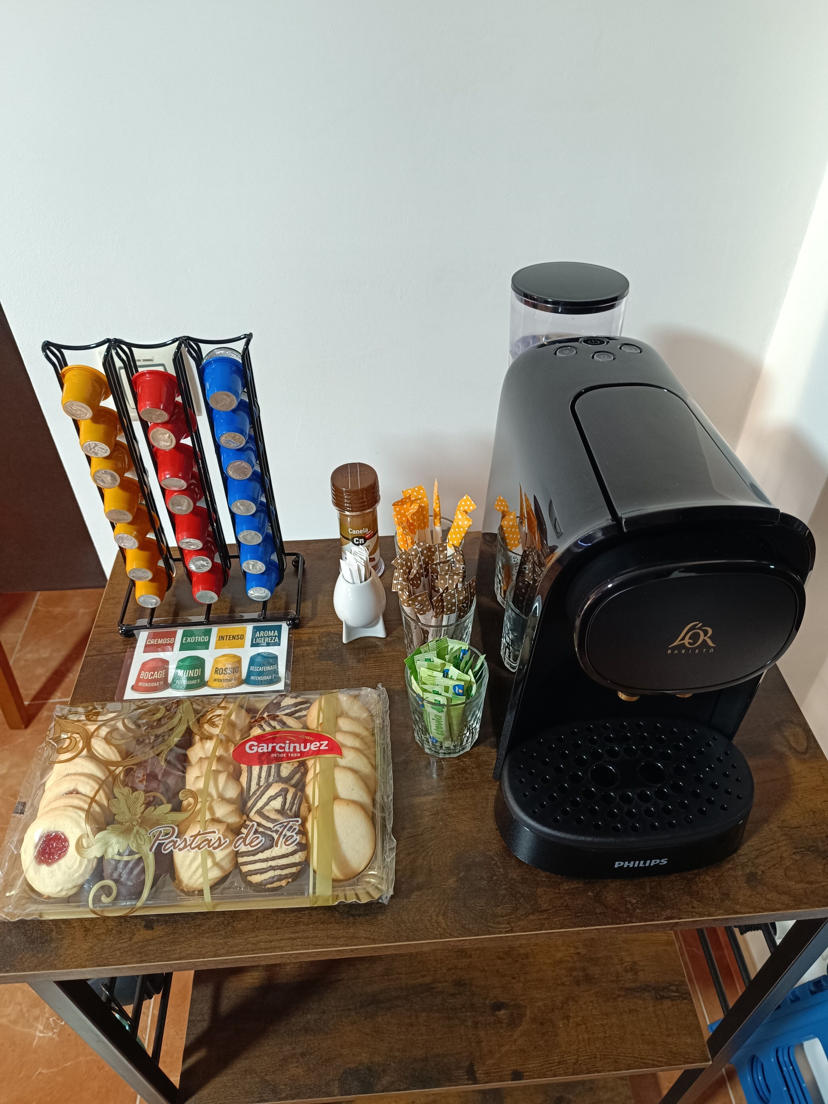 Una cafetera, un tarro de granos de café, un vaso con una cuchara y una caja de galletas sobre una mesa de madera.