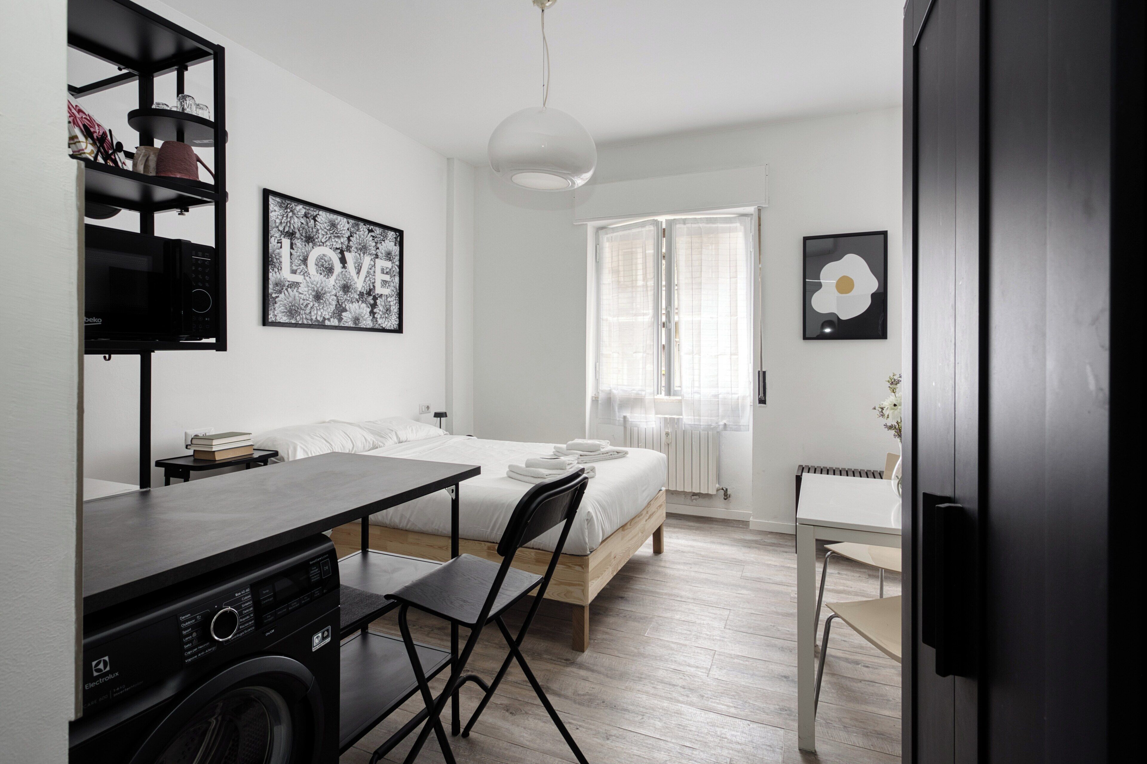A compact hotel room with a bed, a desk, a chair, a washing machine, and a radiator.
