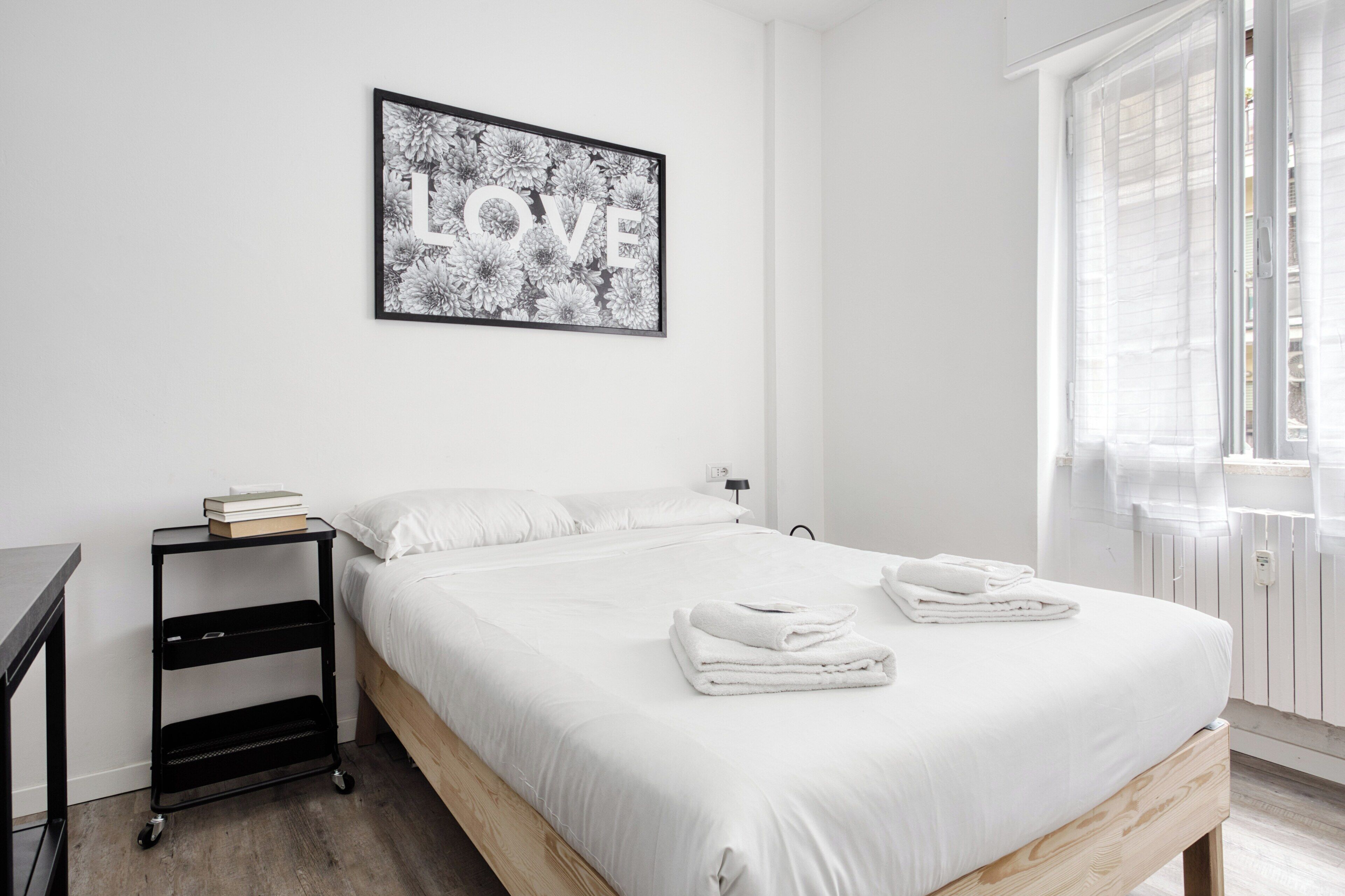 A minimalist bedroom with a bed, a bedside table with books, a framed 'LOVE' artwork, and a window with blinds.