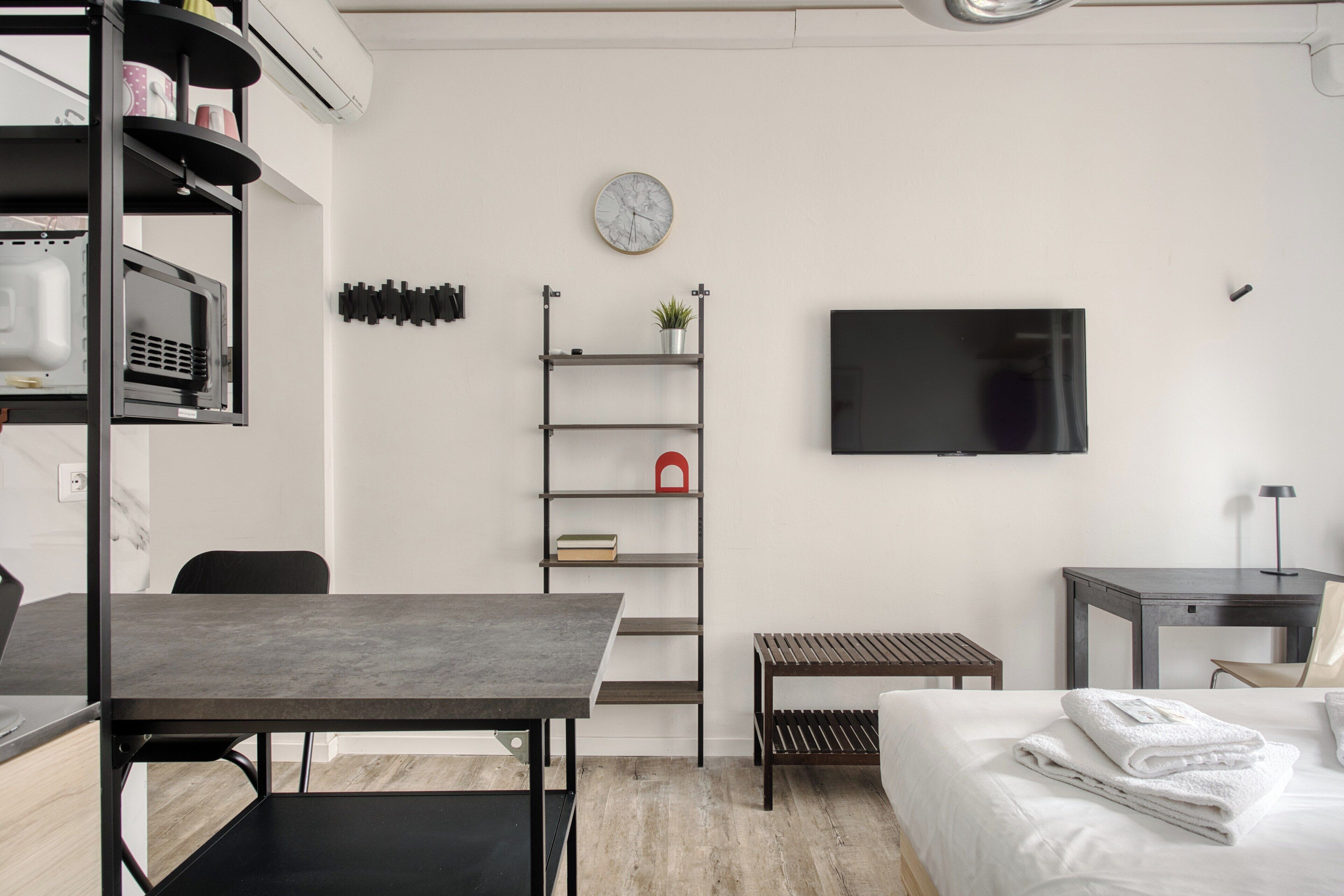 Een moderne hotelkamer met een bed, een bureau met stoel, een televisie, een plank met boeken en een plant, en een klok aan de muur.