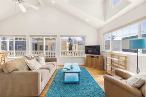 A spacious living room with a beige sofa, a blue coffee table, and a television on a wooden stand. Large windows provide ample natural light and views of the neighborhood. - Capitola Sea Winds one block to Capitola Beach (Capitola)