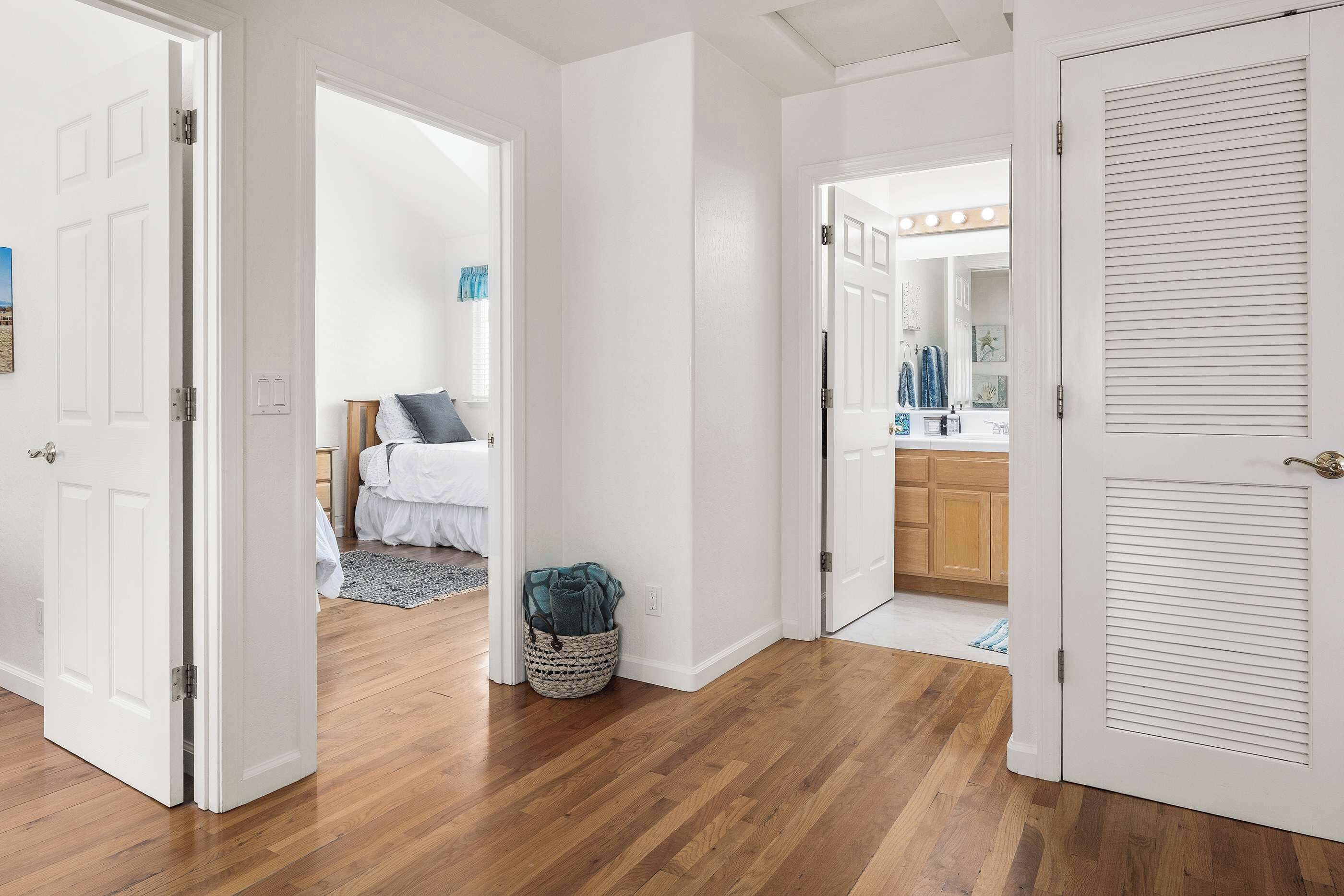 A hallway with wooden flooring, white walls, and open doors leading to a bedroom and a bathroom.