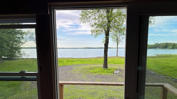 Une vue depuis une pièce dotée d’une grande fenêtre donnant sur un espace verdoyant avec un arbre et une étendue d’eau.