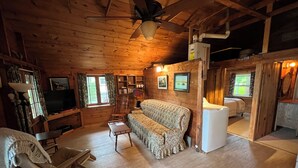 L’intérieur chaleureux d’un chalet avec un plafond en bois, un ventilateur de plafond, un canapé, un fauteuil à bascule, un téléviseur et un réfrigérateur.