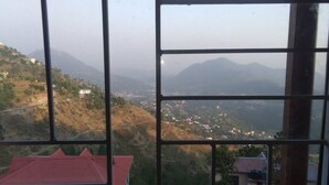 A view through a window with a metal frame, overlooking a hilly landscape with scattered buildings. - Majestic Cottage with WiFi in Bhimtal (Ghorakhal)