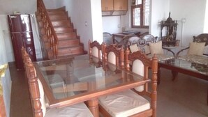 A dining area with a glass-top table and wooden chairs, a staircase, and a kitchen in the background. - Majestic Cottage with WiFi in Bhimtal (Ghorakhal)