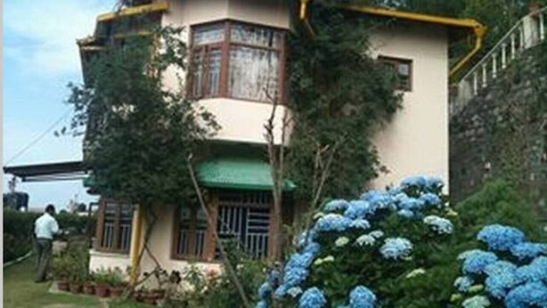 A two-story house with a green roof and a yellow trim. There are blue flowers in the foreground. - Majestic Cottage with WiFi in Bhimtal (Ghorakhal)
