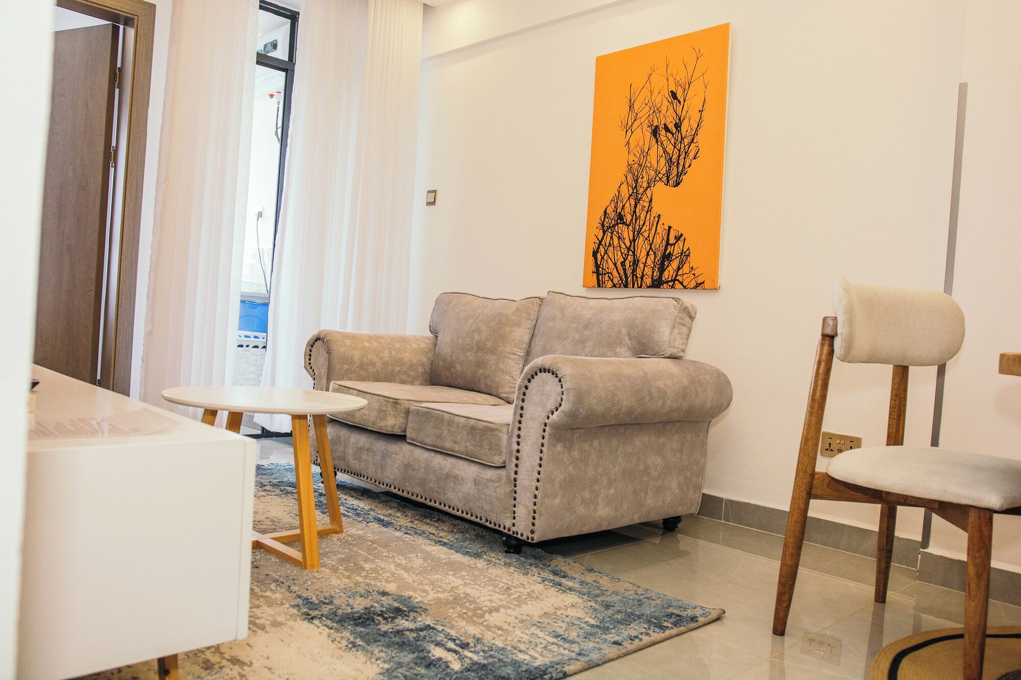 Un salon avec un canapé beige, une chaise en bois, une petite table et un tapis aux motifs bleus et beiges.