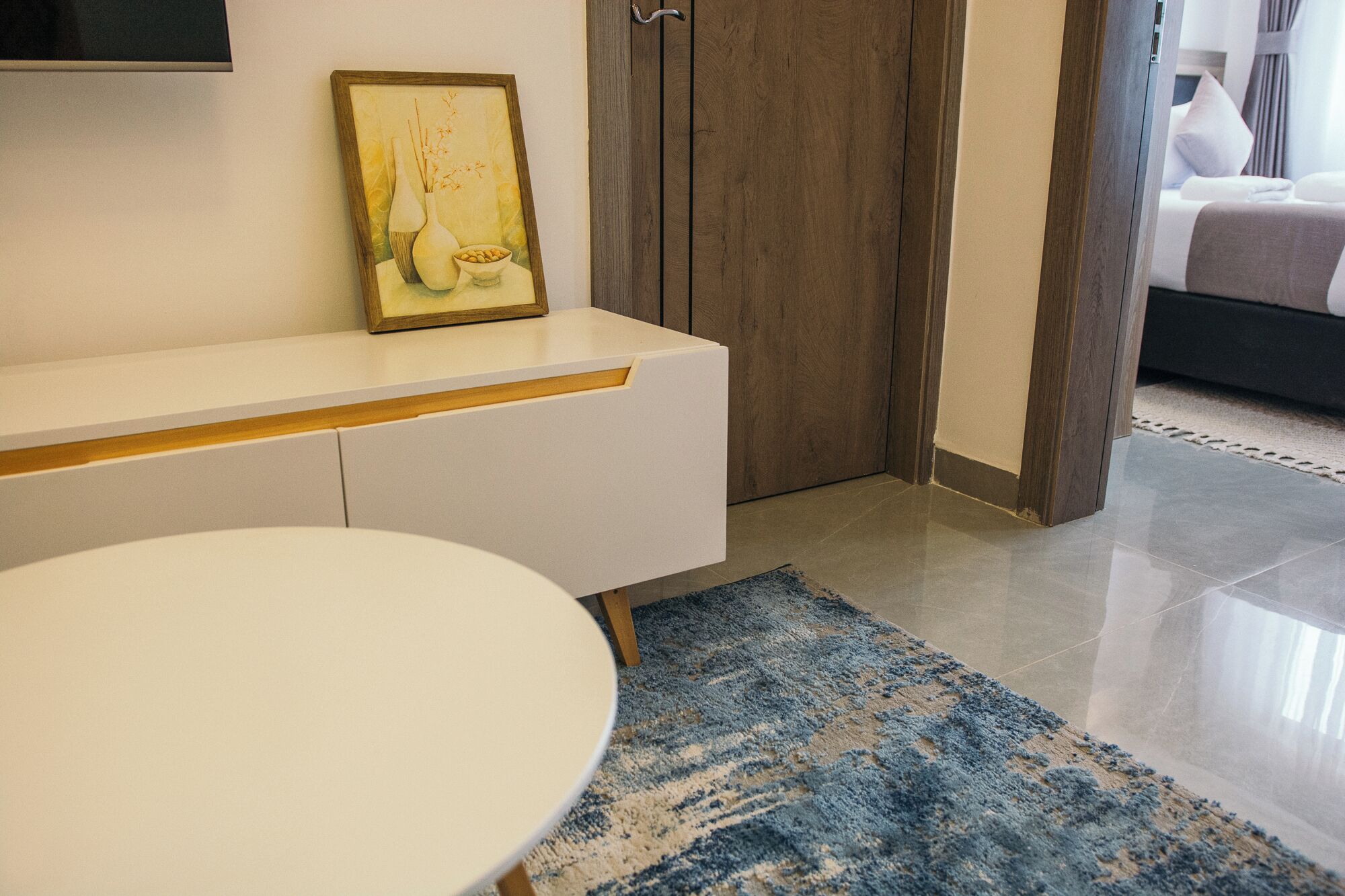 A modern hotel room with a white round table, a blue patterned rug, a wooden door, and a bed with white bedding.