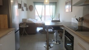 A modern kitchen with a dining area, a bed, and a large window with curtains. - Le Cosy Zen des Sables (Mimizan)