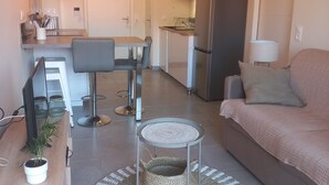 A modern living room with a sofa, a coffee table, and a dining area with a table and chairs. There is a kitchen in the background with a refrigerator and a microwave. - Le Cosy Zen des Sables (Mimizan)