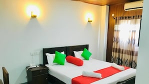 Een net opgemaakt bed met een rood-witte plaid, groene en rode kussens en een witte handdoek. Er staat een nachtkastje met een telefoon en een lamp. De kamer heeft een houten plafond en een raam met gordijnen.