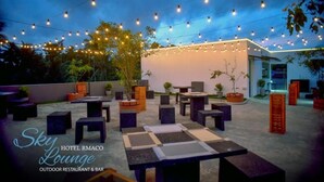 Een terras met tafels en stoelen, sfeerverlichting erboven en planten in potten.