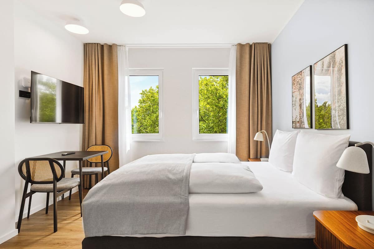 Ein Hotelzimmer mit einem Bett, einem Schreibtisch und zwei mit Vorhängen versehenen Fenstern.