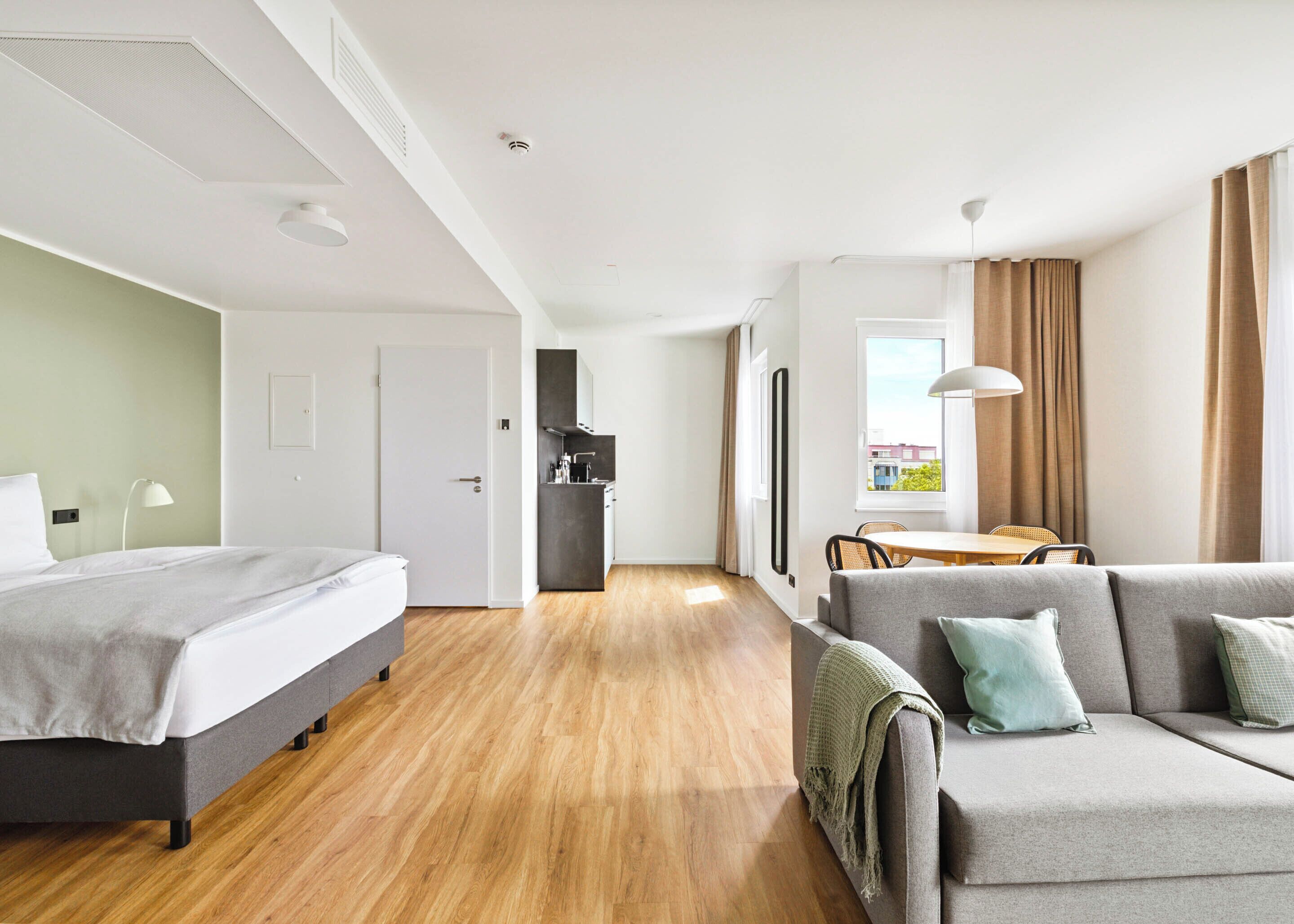 A modern hotel room with a bed, a sofa, a dining area, and a kitchen.