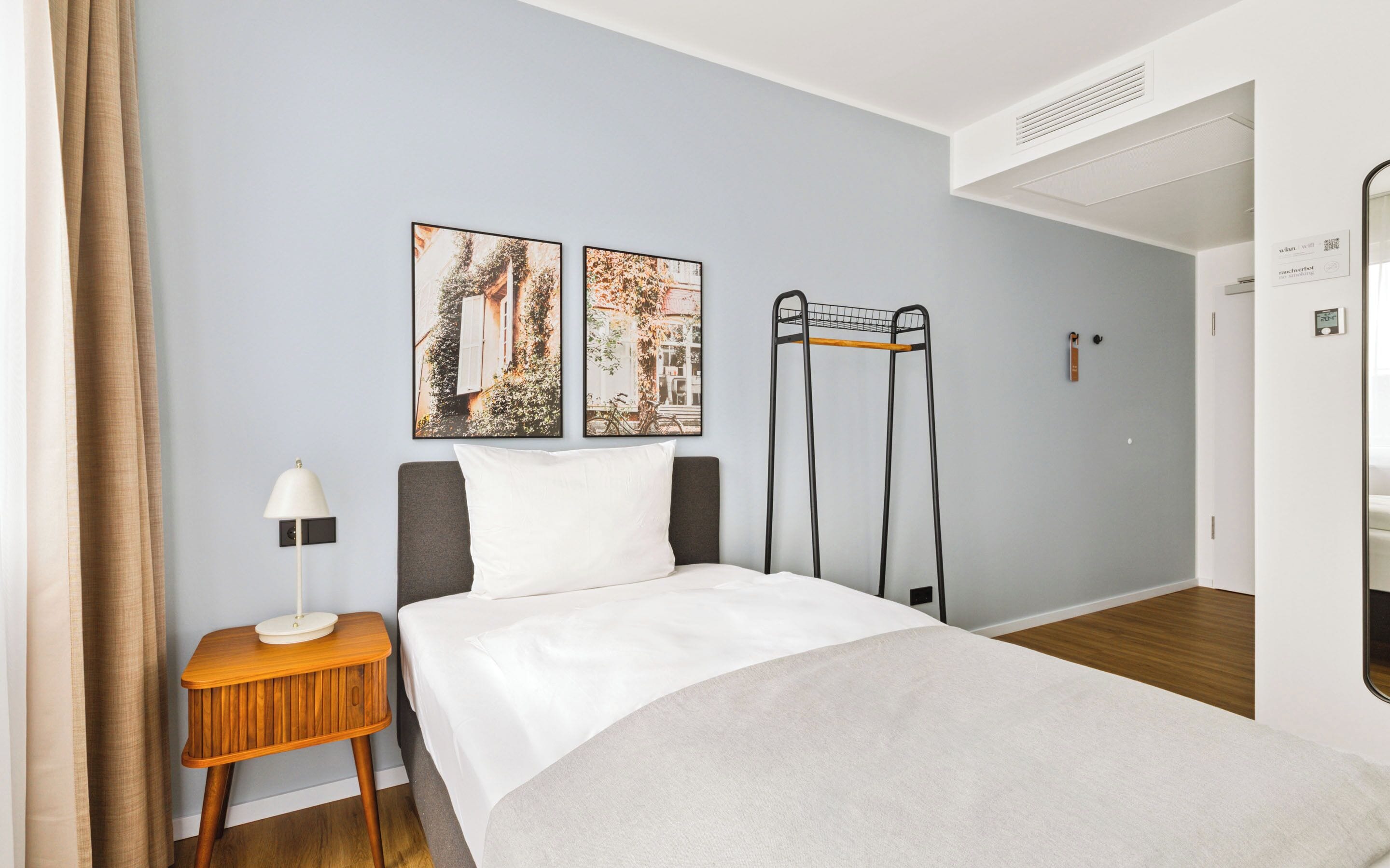 A hotel room with a bed, a nightstand, a lamp, two wall-mounted paintings, a black metal rack, and a door with a control panel.