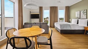 A hotel room with a bed, a sofa, a dining table with chairs, a TV mounted on the wall, and large windows offering a view of the city. - Limehome Bochum Kortumstr. (Bochum)