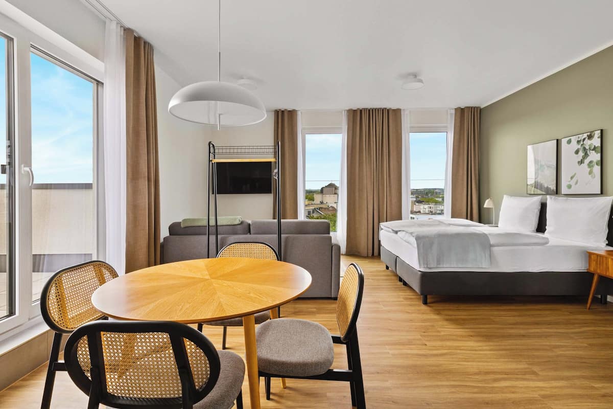 Ein Hotelzimmer mit Bett, einer Couch, einem Esstisch mit Stühlen, einem an der Wand befestigten Fernseher und großen Fenstern mit Blick auf die Stadt.