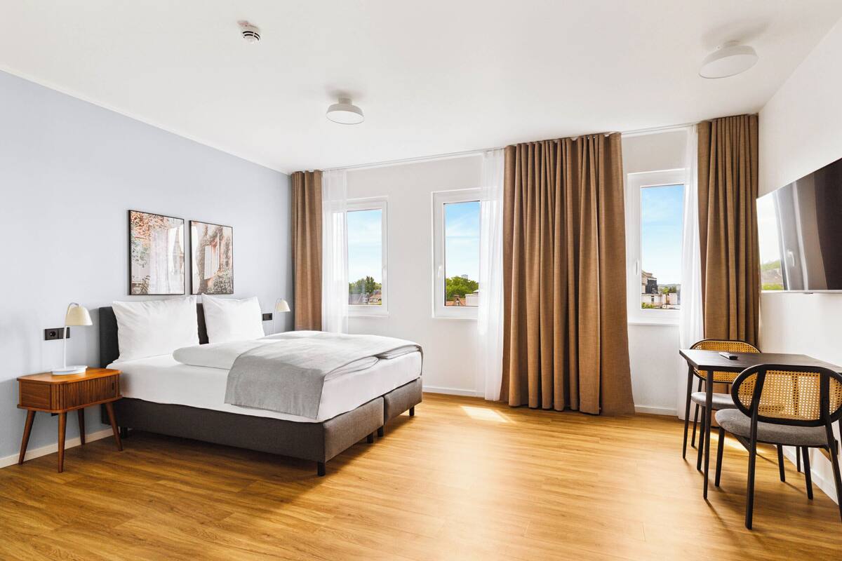 Ein modernes Hotelzimmer mit Bett, Nachttisch, Lampe und Blick auf die Stadt durch große Fenster.