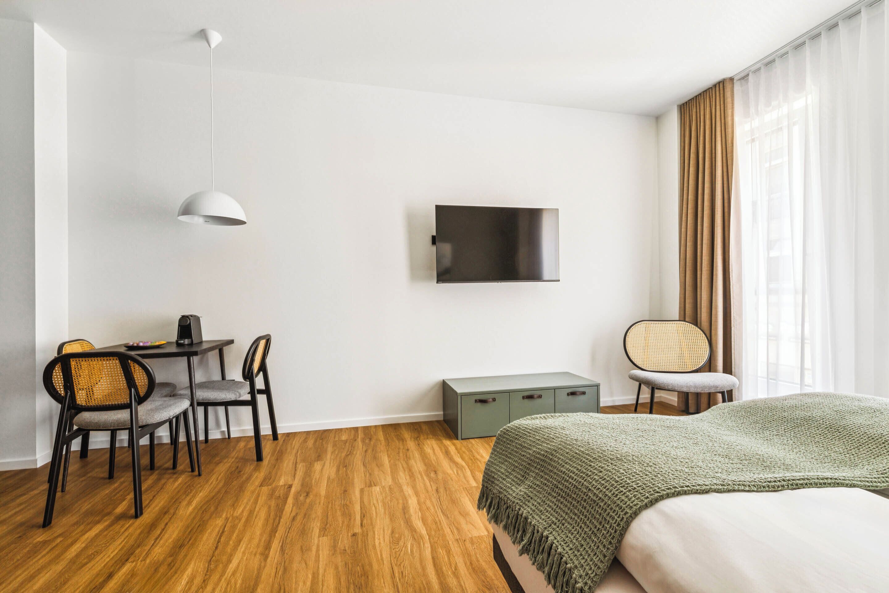 Une chambre d’hôtel moderne avec un plancher en bois, un lit recouvert d’une couverture verte, un bureau avec des chaises, un téléviseur à écran plat fixé au mur et une fenêtre avec des rideaux.