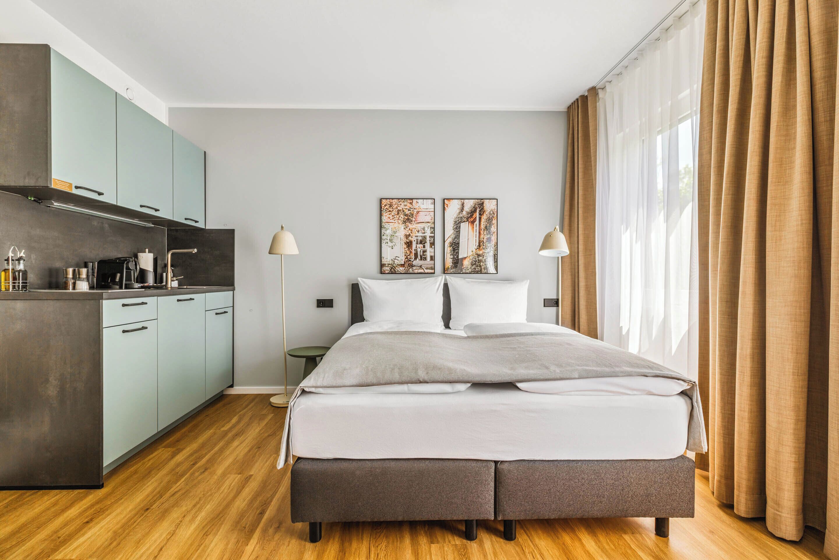 A modern hotel room with a large bed, a kitchenette, and two paintings on the wall.