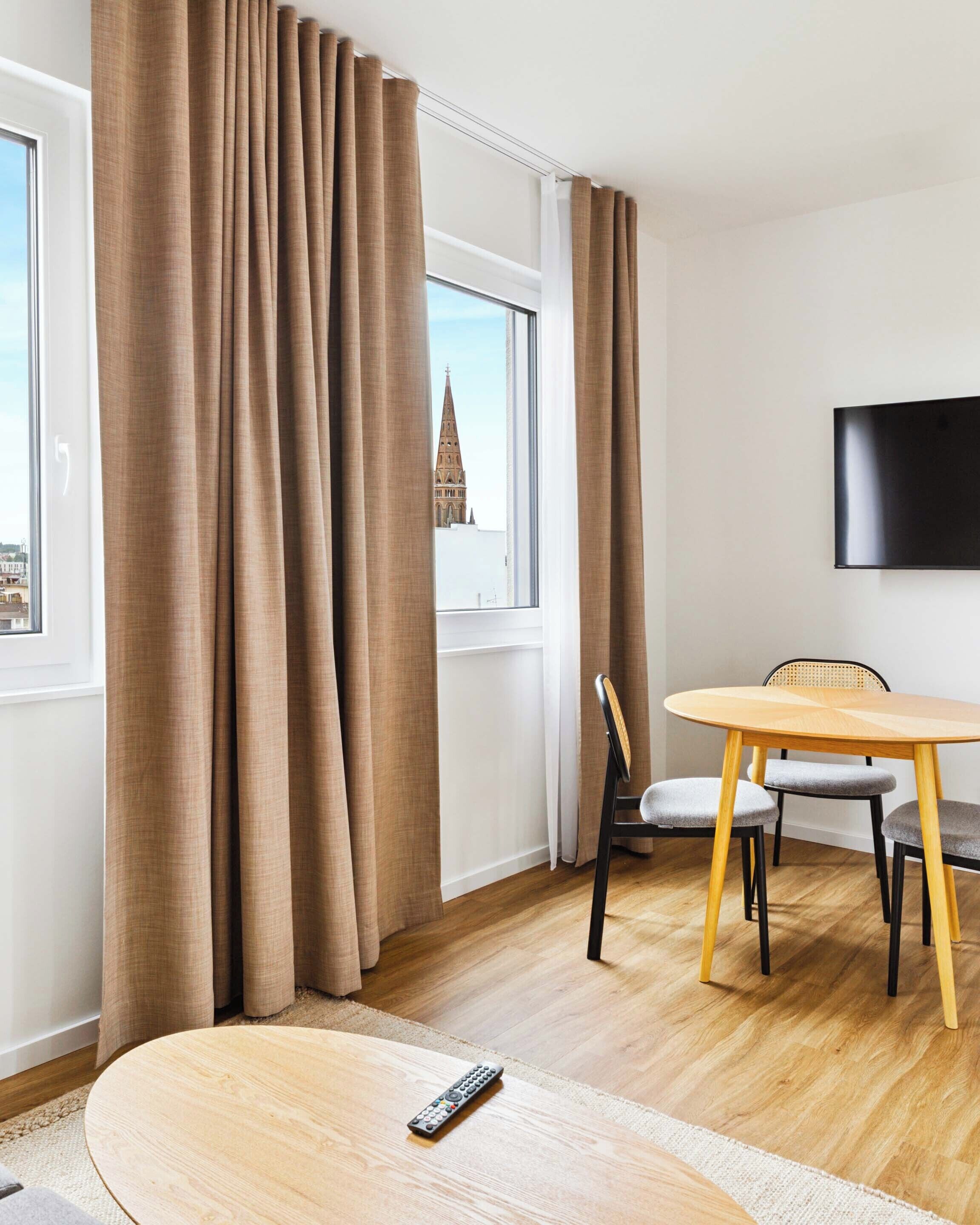 Une chambre d’hôtel moderne dotée d’une grande fenêtre, d’une table ronde en bois, d’un téléviseur fixé au mur et d’une chaise avec un coussin.