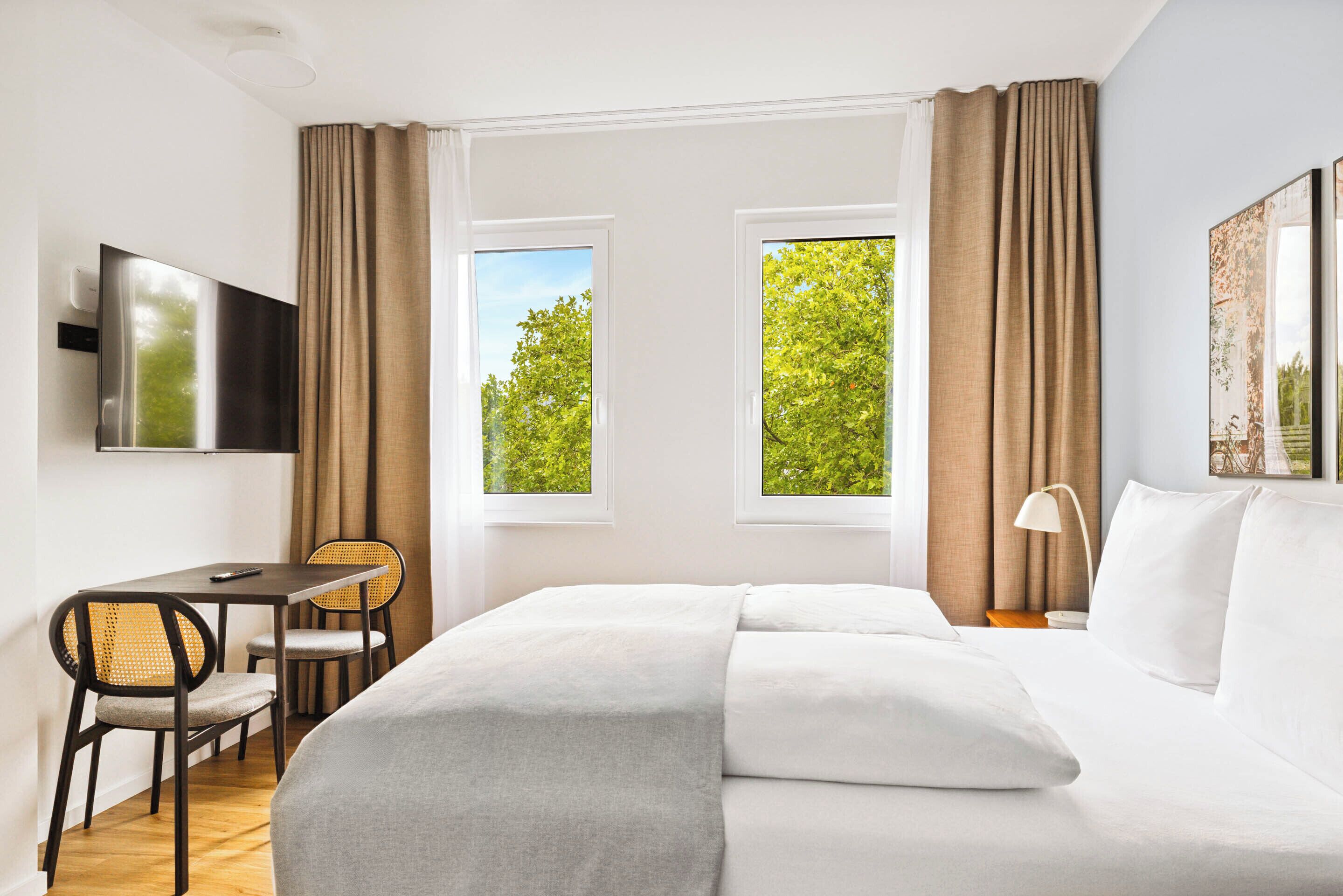 Una habitación de hotel con cama, escritorio y sillas, televisor y dos ventanas con cortinas.