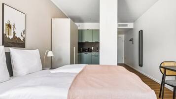 A modern hotel room with a large bed, a bedside table, a lamp, a chair, and a view into the kitchen area.