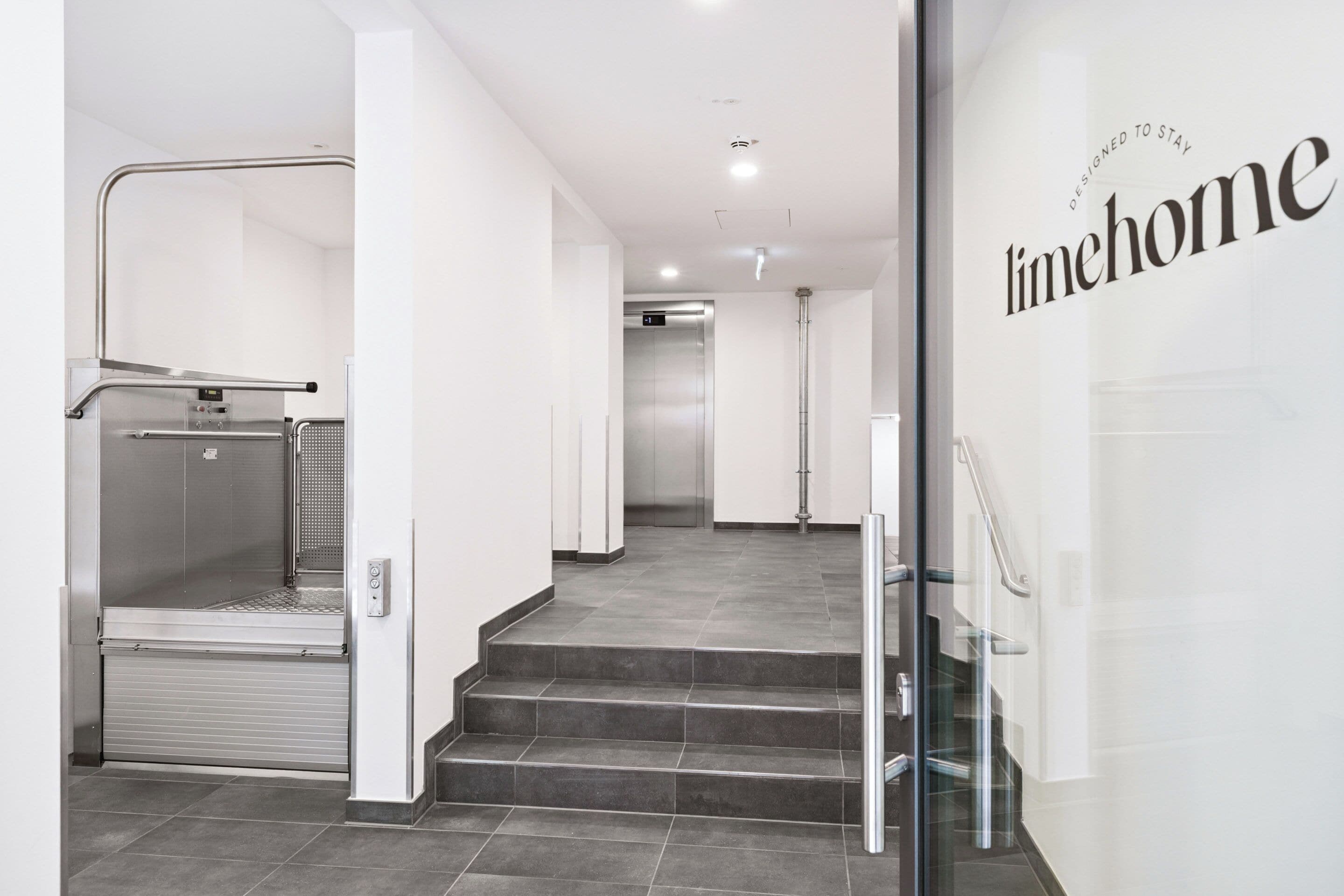 Un interior de hotel moderno con un ascensor de vidrio, una escalera y una pared que muestra el texto 'Designed to Stay'.