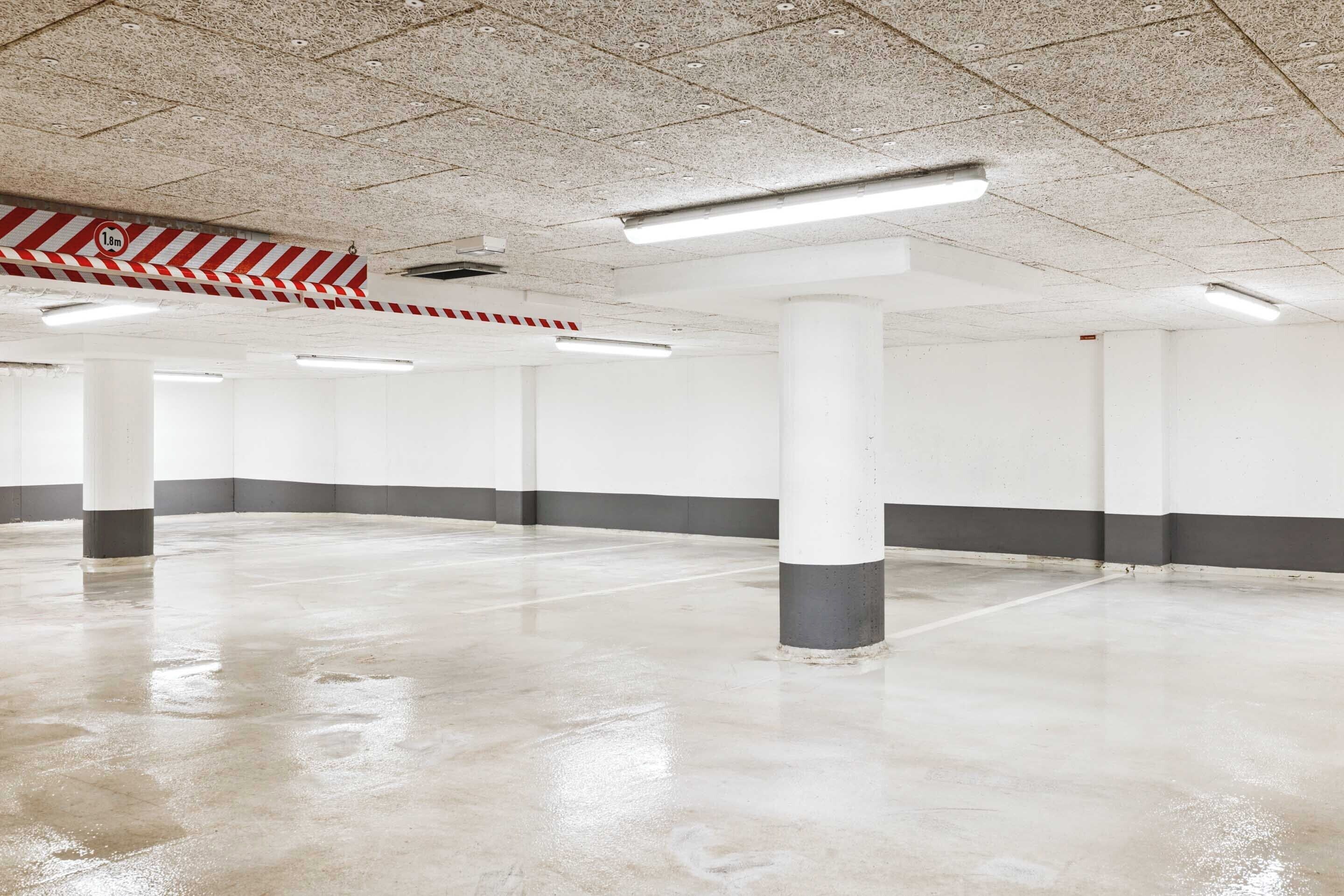A spacious indoor parking area with concrete floors and white walls, featuring several pillars and overhead fluorescent lighting.
