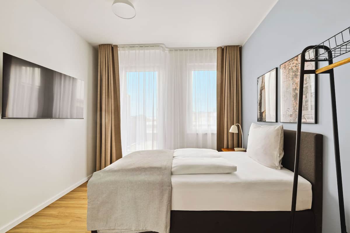 Ein modernes Hotelzimmer mit Bett, Fernseher, Nachttisch mit Lampe und einem Fenster mit Vorhängen.