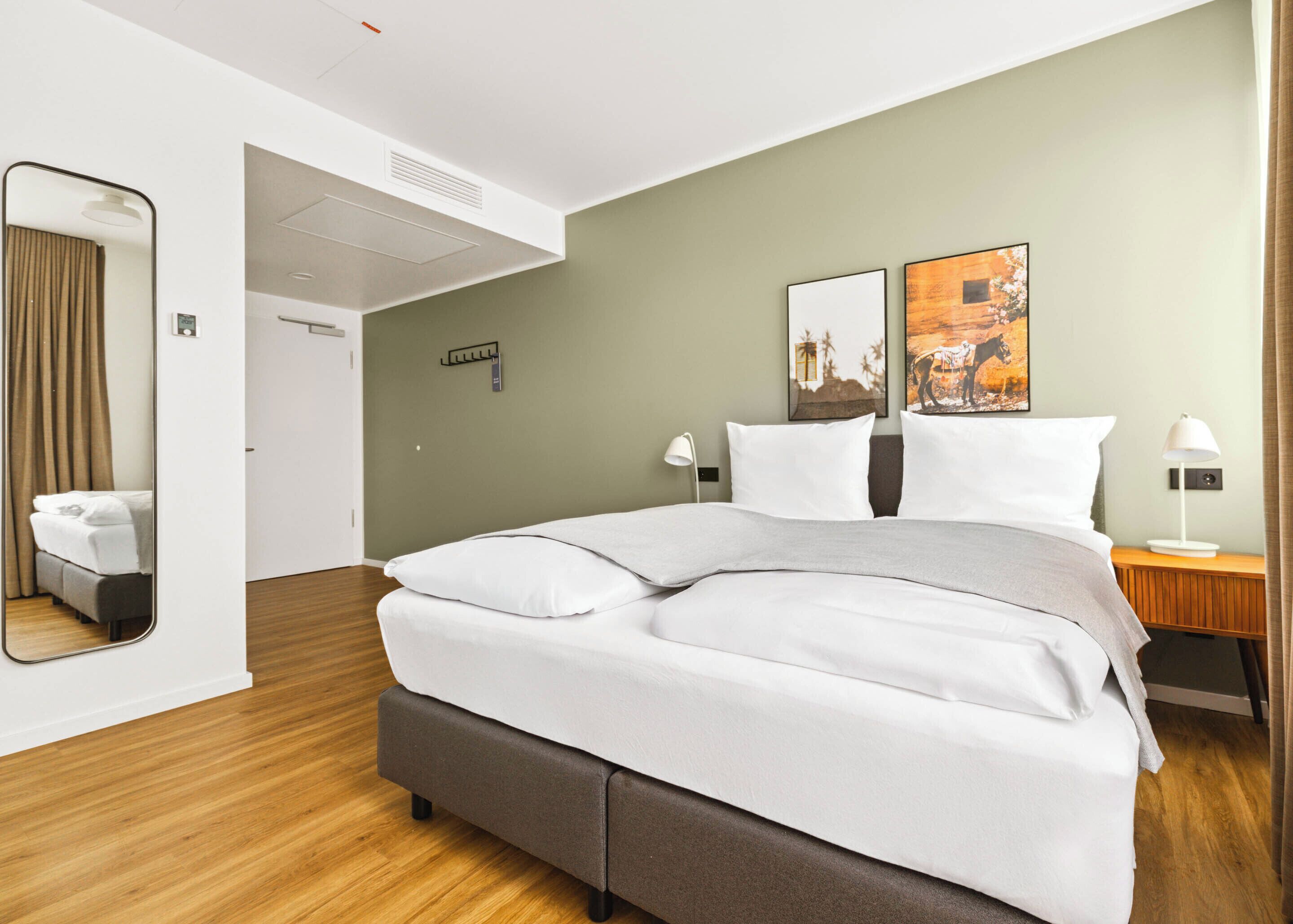 Moderni hotellihuone, jossa on suuri sänky, yöpöytä lampun kanssa, peili ja kaksi seinälle kiinnitettyä taideteosta.