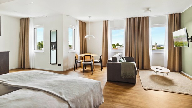 A modern hotel room with a large bed, a sofa, a dining area, and a view of the city. - Limehome Bochum Kortumstr. (Bochum)