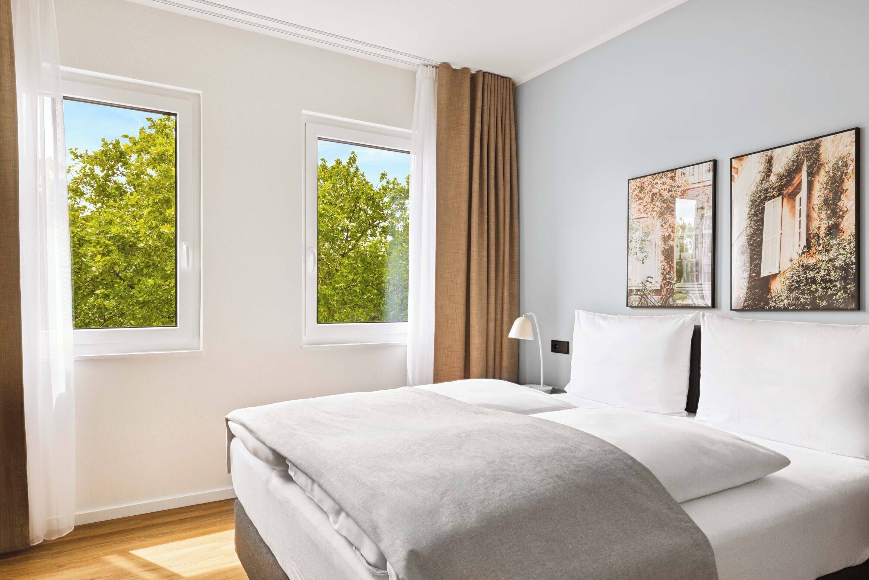 Una camera d'albergo con un letto grande, due finestre, un comodino e due quadri appesi al muro.