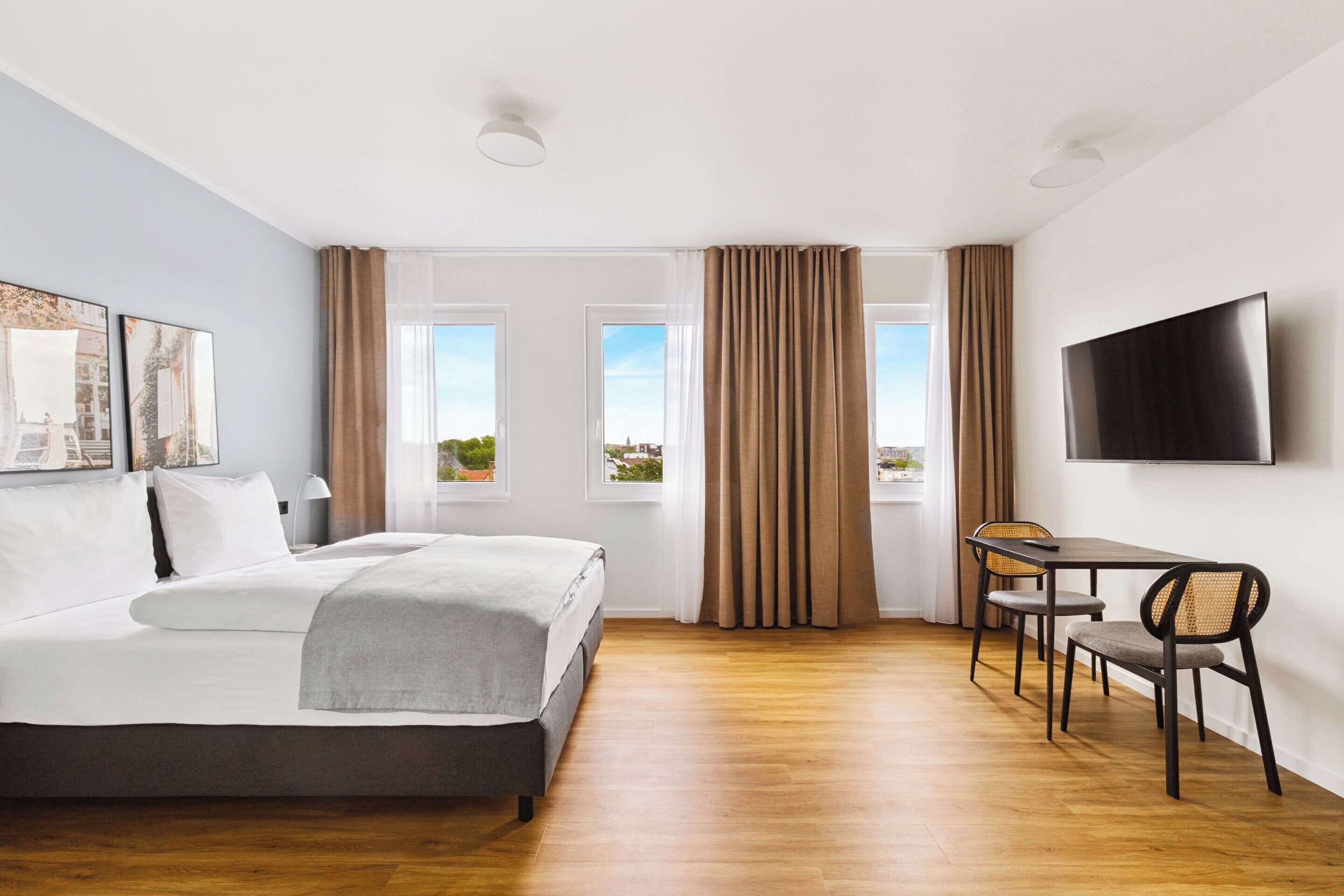 Una habitación de hotel con cama, un escritorio con sillas, televisión y ventanas amplias con cortinas.