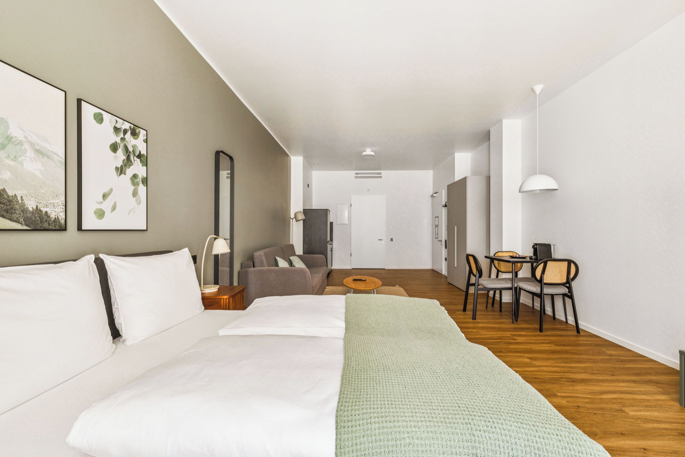A modern hotel room with a large bed, a sofa, a dining area with chairs, and a view of the hallway.