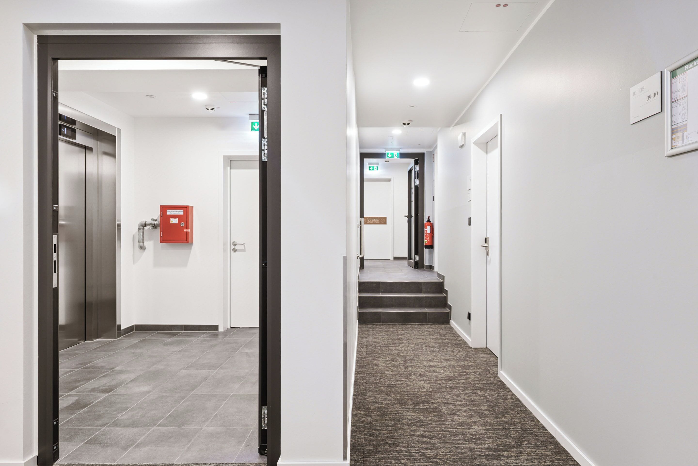 A modern hotel corridor with a carpeted staircase, a fire extinguisher, and an elevator.